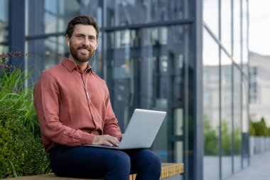 Modern binanın dışında dizüstü bilgisayar kullanan kendine güvenen olgun bir adam profesyonel iş ahlakını gösteriyor ve kentsel yaşam tarzını benimsiyor. Kalabalık şehir hayatının ortasındaki verimliliğe ve bağlantıya odaklan.