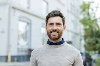 Gülümseyen olgun iş adamı şehir ortamında güvenle dışarıda duruyor. Günlük giysiler içinde, profesyonellik ve dostluk yayıyor..