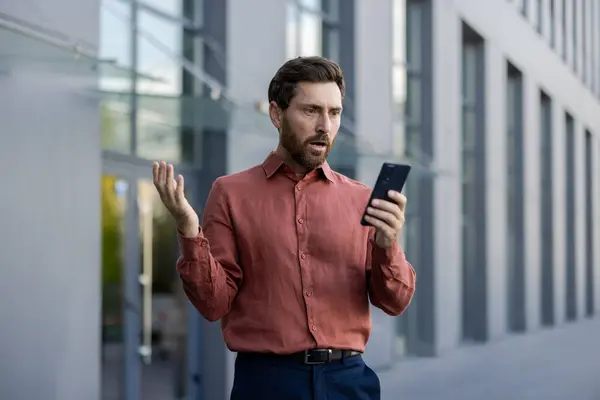 stock image Mature male professional standing outside in city street holding phone, expression of shock and disbelief. Businessman in smart casual attire reacting to surprising news