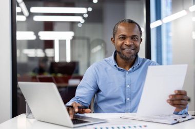 Belgeleri tutarken dizüstü bilgisayar kullanan kendine güvenen bir profesyonel. Modern ofis ortamında olumlu bir tavır sergiliyor. Teknoloji ve evraklar iş analizi öneriyor.
