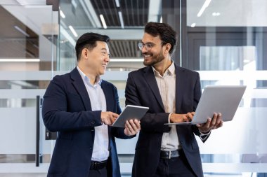 Güvenilir iş adamları tablet ve dizüstü bilgisayar kullanarak dostça sohbet ediyorlar. Görüntü, modern işyeri, takım çalışması ve profesyonel giyimi yakalıyor, işbirliğini ön plana çıkarıyor