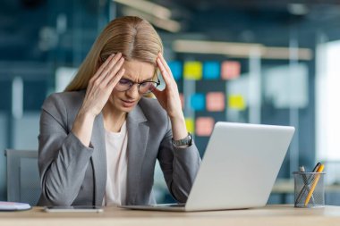 Masadaki gri takım elbiseli iş kadını dizüstü bilgisayarla çalışırken stresli hissediyor. Modern ofis ortamında iş baskısı ve zorluklarla mücadele, hüsran ve endişeyi dile getiriyor.