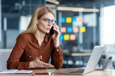 Çağdaş ofiste masa başında çalışırken telefonla konuşan kendine güvenen bir iş kadını. Çalışma günlerinde sohbete, dizüstü bilgisayara ve çoklu görev programlarına odaklanıldı.