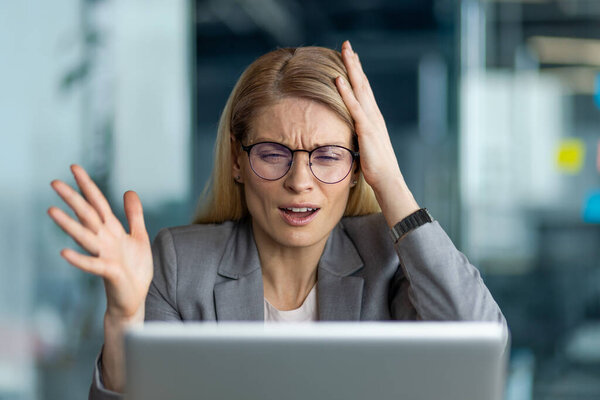 Деловая женщина, чувствующая себя подавленной в офисе, демонстрирует разочарование, указывая на стресс, замешательство и решение проблем в профессиональной обстановке. Женщина в очках и сером костюме отражает проблемы на рабочем месте