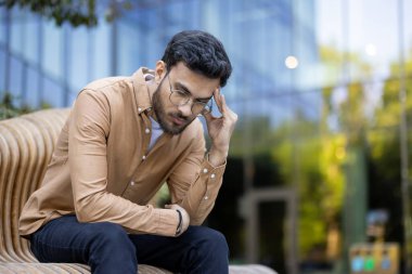 Dışarıda oturan gözlüklü adam stresli ya da derin düşüncelere dalmış görünüyor. O, bir bankta otururken dikkatle odaklanarak günlük giysiler giyiyor. Arka planda modern cam bina görünüyor.