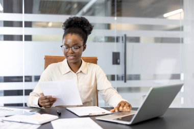 İş kadını büro masasında oturup belgeleri gözden geçirir ve dizüstü bilgisayarla çalışır. Kurumsal ortamda odaklanma ve profesyonelliği gösteren yazılı ve analizli çalışmalar.