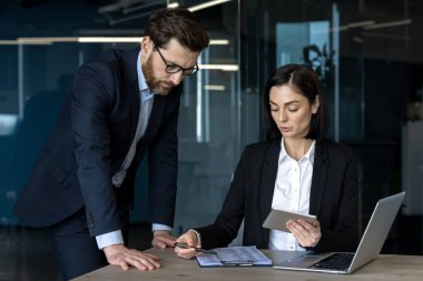İş dünyasından profesyoneller proje detayları üzerinde tartışıyorlar. Kadın referans olarak tablet kullanıyor. Adam masaya eğiliyor, takım çalışmasını vurguluyor. Ofis ortamı teknolojiyi vurguluyor