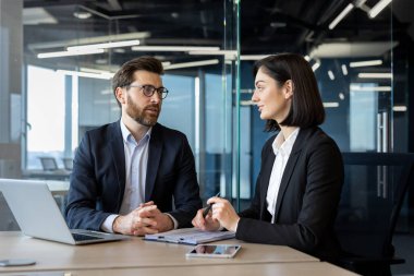 Çağdaş ofiste dizüstü bilgisayar ve belgelerle masada tartışan iki iş adamı. Resmi giyinmiş erkek ve kadın strateji ve işbirliğine odaklanıyor.