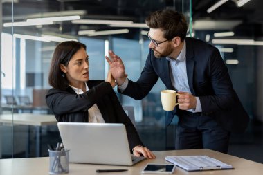 Businesswoman sitting at desk uses strong body language to halt unwanted advance from male colleague with coffee cup, illustrating workplace dynamics, conflict, and harassment prevention. clipart