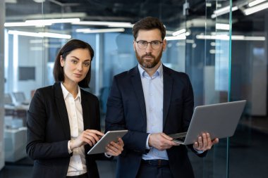 Resmi giyinmiş iş adamları modern ofis ortamında dijital cihazlarla ilgilenirler. Kendine güvenen kadın tableti tutarken, erkek dizüstü bilgisayar kullanıyor, takım çalışması, teknoloji ve yenilik sergiliyor