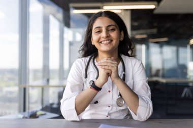 Modern ofis ortamında steteskopla güvenle gülümseyen genç bayan doktor. Görüntü profesyonelliği, bakımı ve yaklaşılabilir davranışı, sağlık temaları için ideal