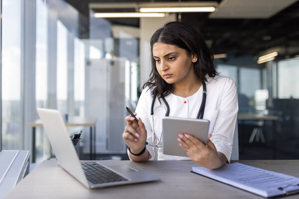 Женщина-врач занимается работой с ноутбуком и планшетом в современном офисе. Основное внимание уделяется медицинским исследованиям и консультированию пациентов. Стетоскоп вокруг шеи указывает на медицинскую профессию.