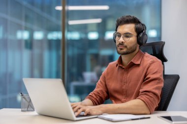 Modern ofis ortamında dizüstü bilgisayarda çalışan kulaklık takan genç bir profesyonel. Günlük kıyafet ve odaklı ifade adanmışlığı ve verimliliği vurguluyor. Uzak çalışma kavramları için ideal