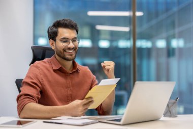 Ofisteki gülümseyen adam zarfı tutuyor, olumlu haberleri heyecanla okuyor. Masada dizüstü bilgisayarla oturuyordu. Başarı ve mutluluk yükseltilmiş yumruk, somut başarı ve motivasyon yoluyla ifade edilir..