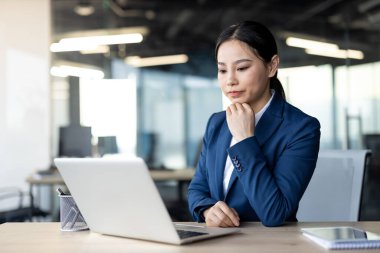 Ofis masasında dizüstü bilgisayar kullanan kendine güvenen Asyalı iş kadını. Kadın işine odaklanmış, profesyonelliği ve konsantrasyonu yansıtıyor. Modern ofis ayarları üretken atmosferi geliştirir.