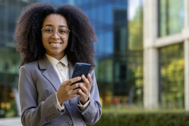 Gülümseyen Afro-Amerikalı iş kadını, modern ofis binasının yanında telefonu dışarıda tutuyor. Mesleki başarı, iletişim ve kentsel iş ortamı güvenini temsil eder.