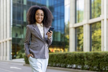 Modern ofis binasının önünde telefonu tutarken gülümseyen kendine güvenen Afro-Amerikan iş kadını. Profesyonel, dengeli ve başarılı kariyer kadını. Liderliği, güçlendirmeyi temsil ediyor.