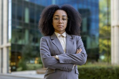 Kendine güvenen Afrikalı Amerikalı iş kadını modern ofis binasının önünde duruyor. Kollarını kavuşturup ciddi bir ifade takınarak profesyonellik sergiliyor.