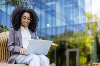 Cam ofis binasının yanında dizüstü bilgisayar kullanan kendine güvenen Afro-Amerikan iş kadını. İş ortamında profesyonel başarı, uzaktan çalışma esnekliği ve modern teknolojiyi temsil eder..
