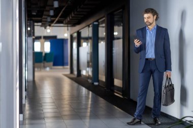 Mavi takım elbiseli iş adamı ofis koridorunda duruyor, akıllı telefonuyla iletişim kuruyor, modern iletişimi gösteriyor. Adam evrak çantasını tutuyor, iş ortamını ve profesyonel kıyafetleri vurguluyor.