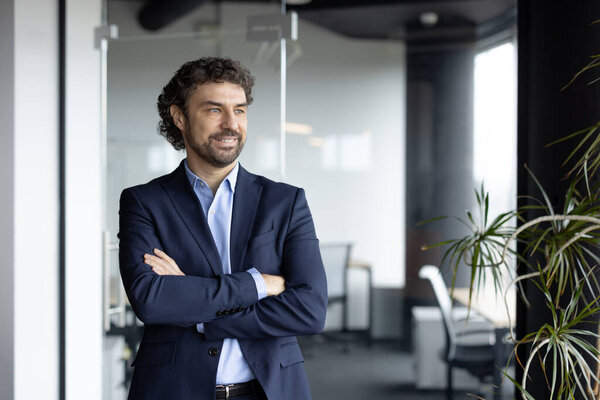 Confident businessman standing in modern office with arms crossed. He appears optimistic and focused, representing success, leadership, and future growth within corporate atmosphere.
