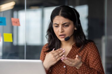 Kulaklık takmış bir kadın, çağdaş ofis ortamında hayal kırıklığına uğramış görünüyor. Yaptığı jestler iş iletişimi sırasında yaşanan yanlış anlaşılmaları ya da kafa karışıklığını vurguluyor.