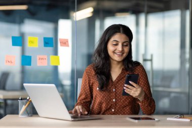 Telefonla gülümseyen ve modern ofiste dizüstü bilgisayarla çalışan bir kadın. Sahne çoklu görev, üretkenlik, iletişim, teknoloji ve iş ortamını temsil ediyor.