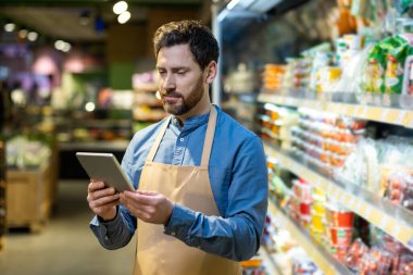 Önlük giyen süpermarket çalışanı market koridorunda tablet kullanıyor. İnsan envanteri modern teknolojiyle yönetir, verimlilik gösterir ve perakende ortamına odaklanır..