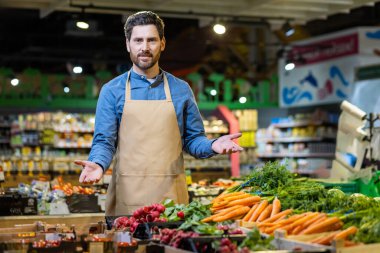 Günlük gömlekli ve önlüklü bir market çalışanı taze ürünlerin arkasında duruyor. Süpermarkette havuç ve turplar düzgünce dizilmiş. Müşteriler için davet atmosferi