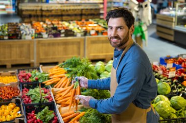 Markette taze sebzeler düzenleyen, havuç tutarken gülümseyen bir market çalışanı. Domates, turp ve marul gibi canlı ürünlerle çevrili. Sağlıklı beslenmeyi temsil eder.