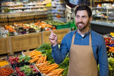 Önlük takan bir market çalışanı taze ürünler bölümüne işaret ederken gülümsüyor. Sahne, canlı süpermarketlerde havuç, turp, yeşillik gibi çeşitli sebzeleri yakalıyor..