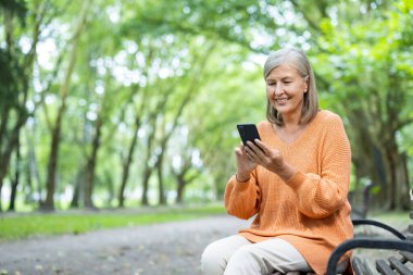 Park bankında oturan son sınıf öğrencisi telefon kullanıyor, turuncu bir kazak giyiyor, gülümsüyor ve dışarıda huzurlu bir günün tadını çıkarıyor, rahatlama ve doğa ve teknolojiyle olan bağlantısını gösteriyor..