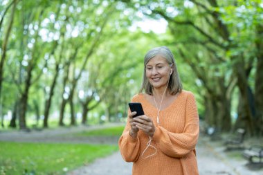 Parkta yürürken akıllı telefondan müzik dinleyen gülümseyen olgun bir kadın. Kulaklık takmak, rahatlamak ve memnun olmak. Keyif ve sükuneti yansıtan doğal ortam.