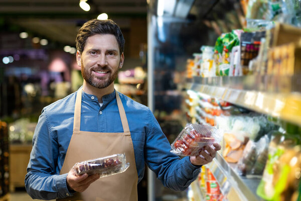 Smiling grocery store employee holding fresh produce in a supermarket aisle. Displays friendliness and quality selection, emphasizing customer service. Surrounded by an array of products