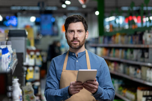Supermarket employee wearing apron stands in grocery aisle holding digital tablet. Shelves lined with products present busy retail environment. Man appears focused, embodying themes of retail