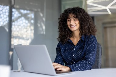 İspanyol iş kadını masada dizüstü bilgisayar kullanarak oturuyor. Puantiyeli tişört giyiyor ve gülümsüyor. Olumlu ve üretken bir atmosfer yaratıyor..