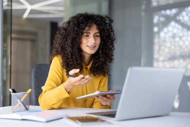 Çevrimiçi toplantı yapan kendine güvenen İspanyol iş kadını. Masaya oturmuş, dizüstü bilgisayar kullanıyor ve not defteri tutuyor. Profesyonelliği, iletişimi, uzaktan çalışmayı temsil eder.