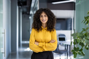 İspanyol iş kadını modern ofiste kollarını kavuşturarak rahatça ayakta duruyor. Gülümsüyor, profesyonelliğini ve kararlılığını gösteriyor. Şirket ortamında teknoloji ipuçları içeren ofis ayarları.