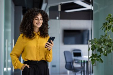 İspanyol iş kadını ofiste telefonlarını kontrol ederken gülümsüyor. Sarı bluz giyiyor, özgüven ve profesyonellik sergiliyor, modern iletişimle meşgul..