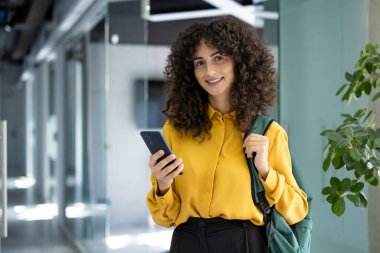 Modern koridorda sarı tişört ve sırt çantası giyen İspanyol öğrenci elinde kendine güvenen bir gülümsemeyle akıllı telefon tutuyor. Resim eğitim, teknoloji ve çağdaş yaşam tarzı temaları taşır.