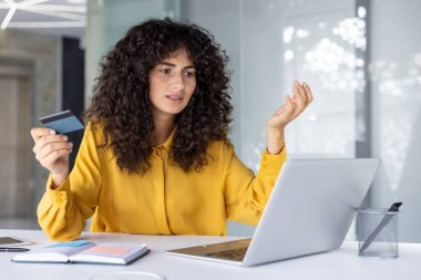 Hüsrana uğramış bir ifadeyle dizüstü bilgisayara bakarken elinde kredi kartı olan bir kadın. Çevrimiçi ödeme sorunu veya olası dolandırıcılık, işlem veya hesap hatası üzerine kafa karışıklığını gösterir.