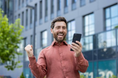 Bir adam cep telefonu tutuyor ve gülümsüyor. Bir binanın önünde duruyor..