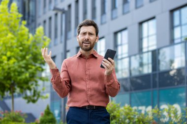 Kırmızı gömlekli bir adam cep telefonu tutuyor. Telefona bakıyor ve çok üzgün.