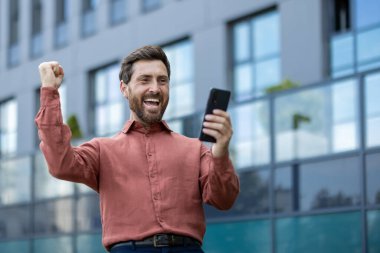 Bir adam cep telefonu tutuyor ve gülümsüyor. O kırmızı bir gömlek giyiyor. Arkasındaki bina gri.