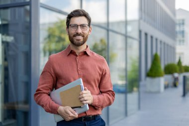 Gözlüklü ve kırmızı gömlekli bir adam bir dizüstü bilgisayar ve bir kağıt parçası tutuyor. Gülümsüyor ve bir binanın önünde duruyor.