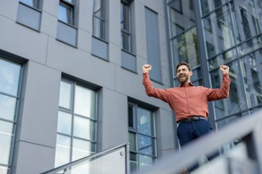 Turuncu gömlekli bir adam balkonda duruyor ve ellerini havaya kaldırıyor. Gülümsüyor ve mutlu.