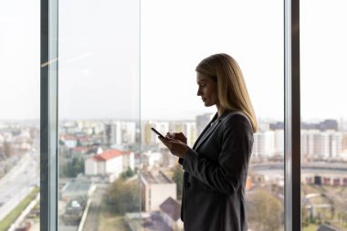 Ofis penceresinin yanında akıllı telefon kullanan resmi giyinmiş bir iş kadını. Parlak gün ışığı profesyonel atmosferi güçlendirir. Şehir manzarası dijital iletişim odaklı kentsel ayarı öneriyor.