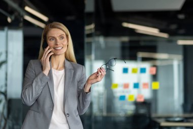 Gri takım elbiseli kendine güvenen kadın ofisinde elinde gözlükle akıllı telefondan konuşuyor. İş iletişimini, çoklu görevi, modern profesyoneli sembolize eder. Parlak ofis verimli bir ortam belirtiyor.