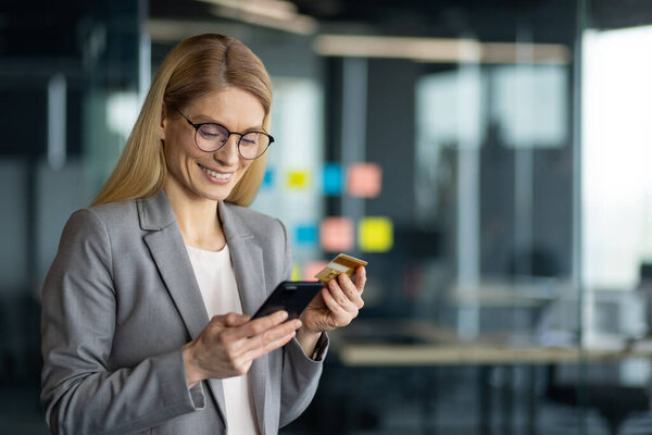 Smiling businesswoman using smartphone for online shopping with credit card in office setting. Professional attire, modern technology, and confident demeanor emphasize efficiency and connectivity.