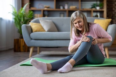 Diz ağrısı olan yaşlı bir kadın evde egzersiz yaparken yerde oturuyor ve rahatsızlığını ve endişelerini dile getiriyor. Oturma odasında dinleniyor, yeşil yoga minderinde dinleniyor, günlük giysiler giyiyor..